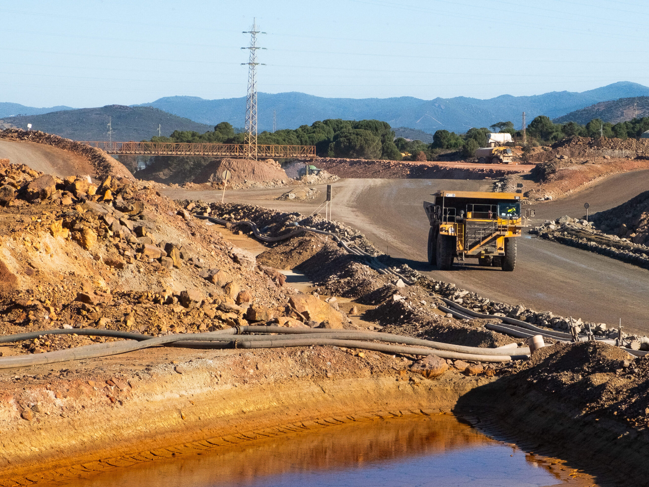 Scène d'exploration minière dans un paysage de collines verdoyantes. Un grand camion-benne jaune circule sur une rampe d'accès en terre. Au premier plan, un bassin d'eau de drainage minier acide de couleur orange vif, entouré de tuyaux noirs. Au loin, des pylônes électriques et des massifs montagneux sous un ciel clair.